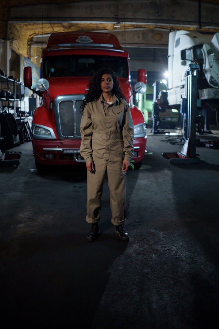 Woman in mechanic uniform standing confidently in an auto repair shop with a truck behind.